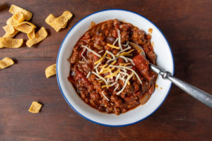 bowl of chili with beans and tomatoes topped with shredded cheese with side of corn chips flat lay